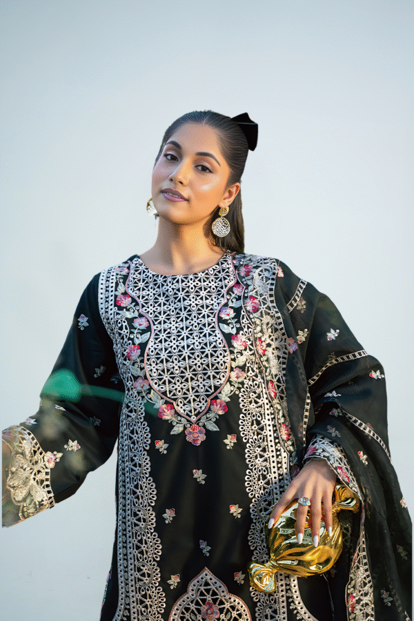 Woman wearing an elegant black embroidered long dress with beige and pink floral detailing, styled with gold clutch and pink heels