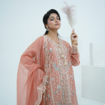 A model wearing a peach embroidered lawn Eid dress with intricate boring design and organza border, paired with a cotton net dupatta with embroidered borders – Amzoclothing festive collection.